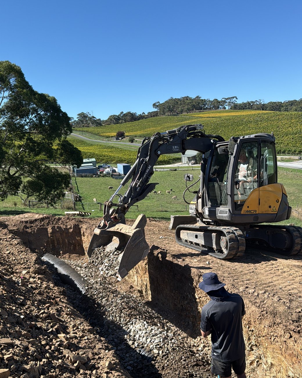 An excavator digging a gravel-lined drainage trench on a hillside job, with vineyards behind
